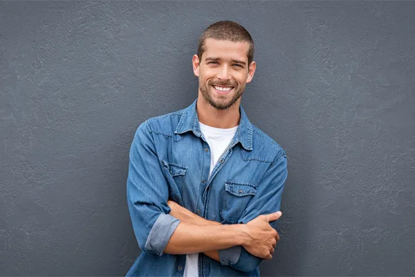 Hormones-for-Men-Doctor A man in a denim shirt stands smiling against a gray-blue wall, pleased with his testosterone hormone therapy from Fonthealth in Niagara.