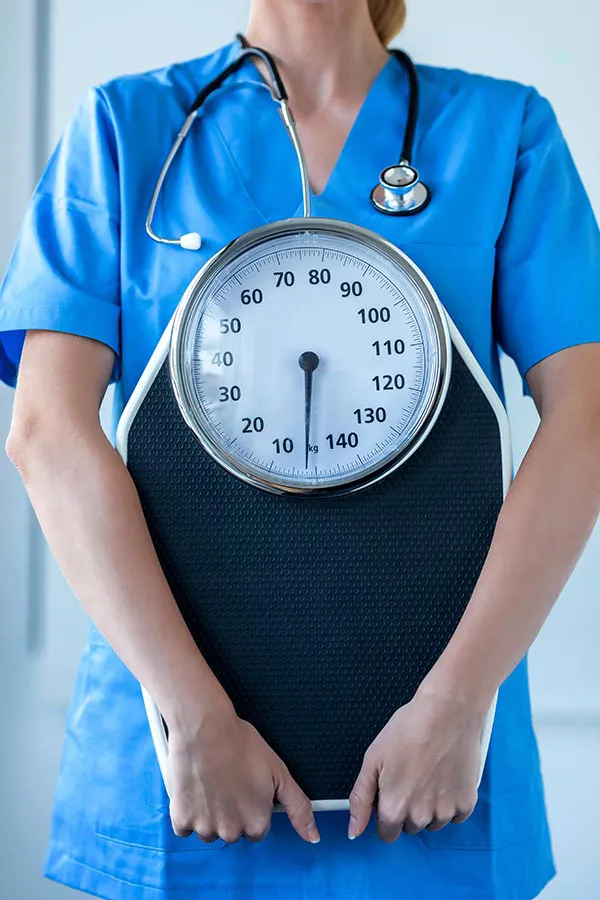 Medical-Weight-Loss-Treatment A nurse in blue scrubs with a stethoscope hanging around her neck holds a scale, representing weight loss treatments from Fonthealth in Niagara.