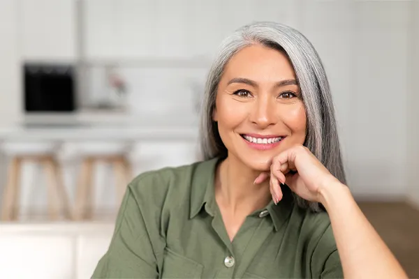 Menopause-Clinic An asian woman with long gray hair and a green shirt sitting and smiling on her couch, happy with her menopause care from [NAME} in Niagara.