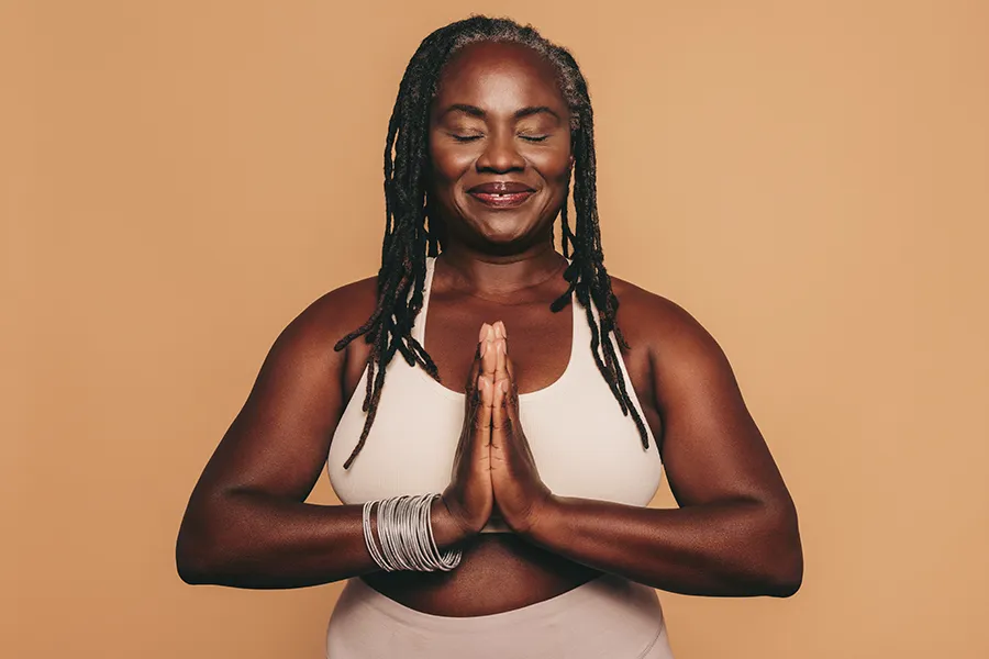 Obesity-Clinic An older African American woman confidently posing against a warm background smiling with hands in a prayer pose. Fonthealth treats obesity using functional medicine in Niagara.