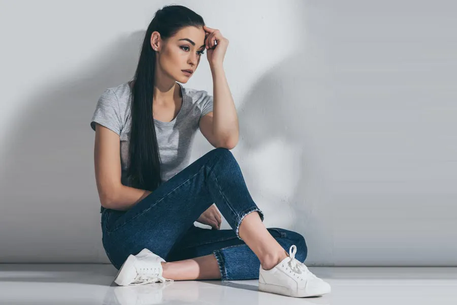 PCOS-Doctor A young brunette woman in a gray t-shirt and jeans sits against a gray wall with her head resting on her hand and a distressed expression. Get help for Polycystic Ovarian Syndrome (PCOS) from Fonthealth in Niagara.
