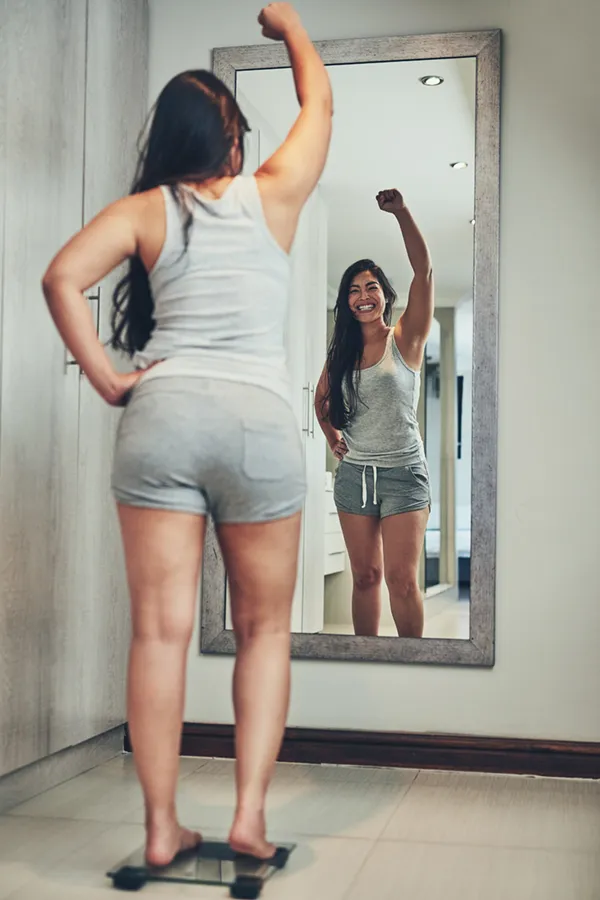 Tirzepatide-Treatment A woman in workout clothes stands on a scale looking in the mirror and pumping her hands in celebration of her successful Tirzepatide treatment from Fonthealth in Niagara.
