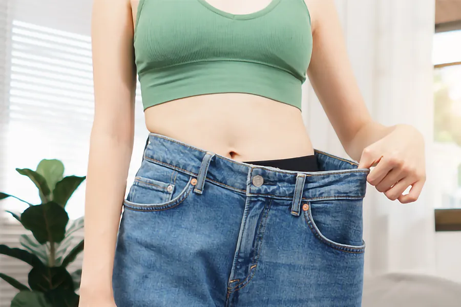 Zepbound-Weight-Loss-Clinic A close-up of a young woman in a green crop top, holding out her oversized blue jeans to show significant weight loss. Zepbound Weight Loss Injections offered by Fonthealth in Niagara.